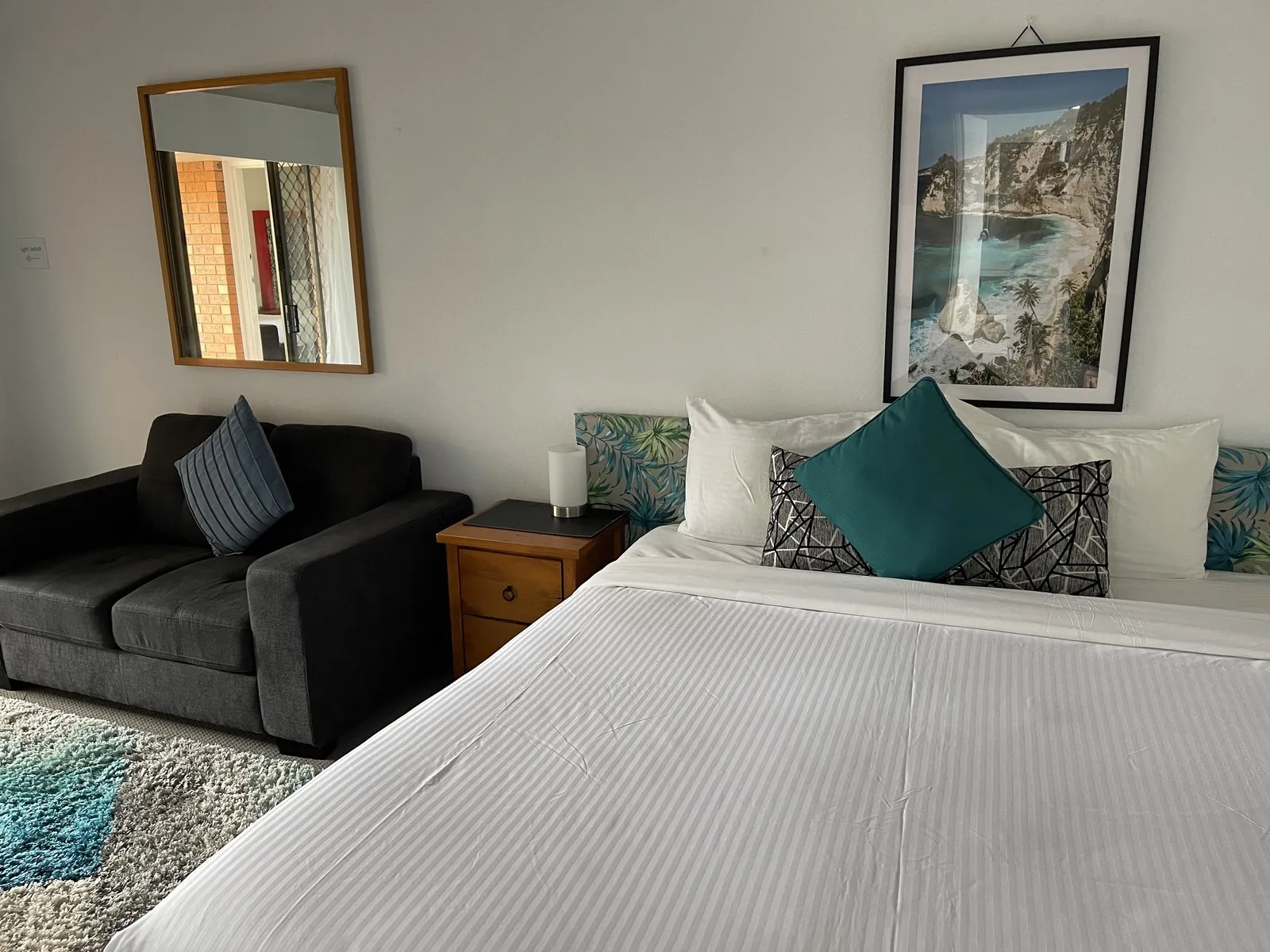 A king room with crisp white linen, teal cushions, a grey sofa and a framed coastal print